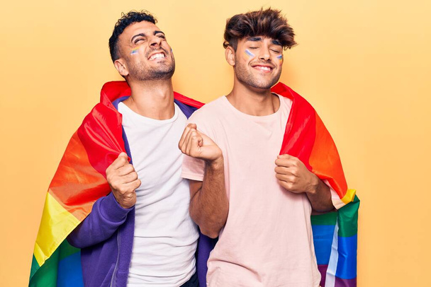 Junges schwules Paar trägt Regenbogenfahne und schreit stolz, feiert Sieg und Erfolg sehr aufgeregt mit erhobenem Arm  - Foto, Bild