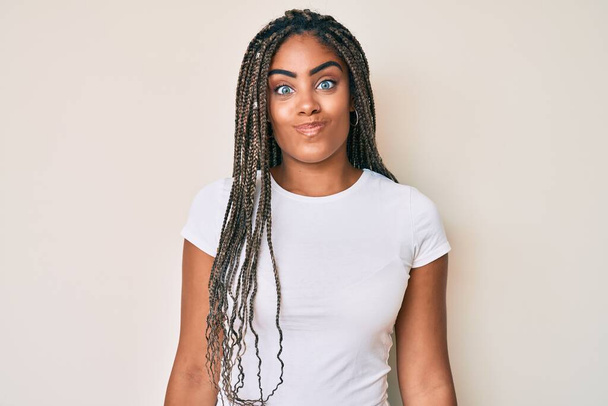 Молодая африканская американка с косичками в обычной белой футболке, пыхтящей щеками с забавным лицом. рот надутый воздухом, сумасшедшее выражение.  - Фото, изображение