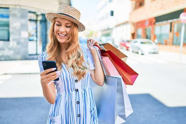 Genç, güzel alışverişçi kadın gülümseyerek markete gidiyor alışveriş torbaları tutuyor, akıllı telefon kullanarak mutlu bir şekilde gülümsüyor. - Fotoğraf, Görsel