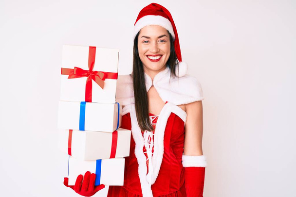 Noel Baba kostümü giyen genç bir kadın elinde hediyeler tutarken pozitif ve mutlu görünüyor ve kendine güvenen bir gülümsemeyle dişlerini gösteriyor.  - Fotoğraf, Görsel