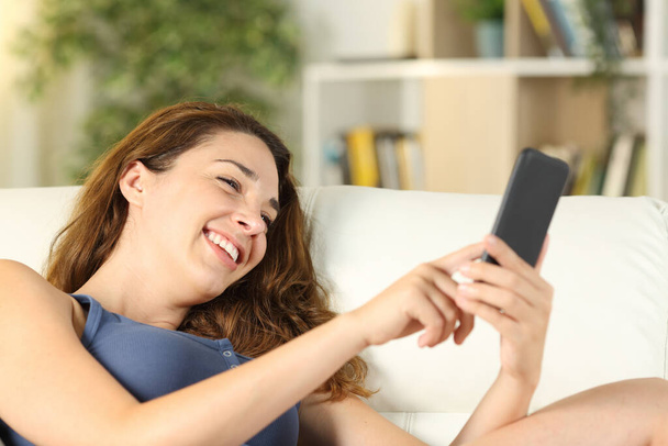 Happy woman using smart phone lying on a couch in the living room at home - 写真・画像