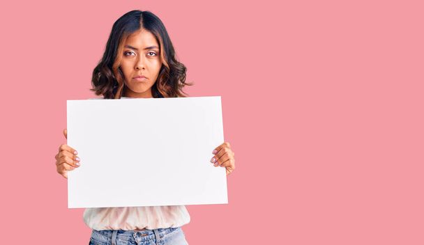 Young beautiful mixed race woman holding cardboard banner with blank space thinking attitude and sober expression looking self confident  - Photo, image