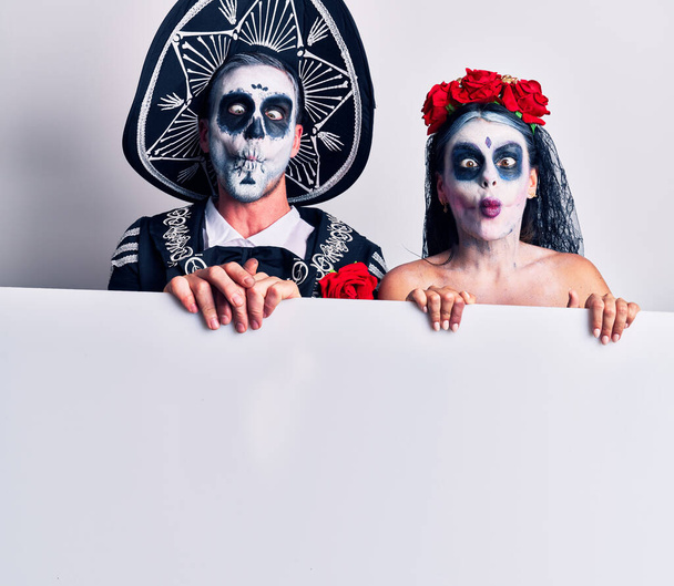 Молодая пара, одетая в мексиканский день мертвого костюма, держит пустой баннер, делая рыбное лицо с губами, сумасшедшим и смешным жестом. Смешное выражение.  - Фото, изображение