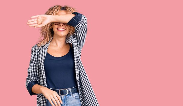 Young blonde woman with curly hair wearing business jacket and glasses covering eyes with arm smiling cheerful and funny. blind concept.  - Photo, Image
