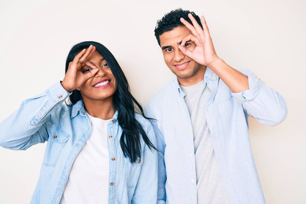 Bella latina giovane coppia indossa abiti casual facendo gesto ok con la mano sorridente, occhio guardando attraverso le dita con il viso felice.  - Foto, immagini