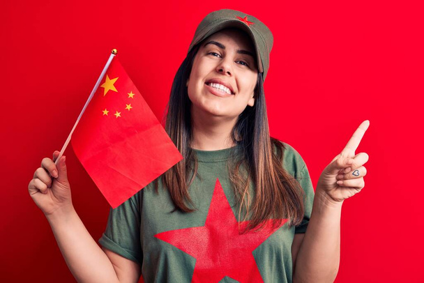 Gyönyörű hazafias nő, piros csillagos kommunista szimbólumos pólóban, porcelánzászlóval a kezében, boldog mosollyal, kezével és ujjával az oldalán. - Fotó, kép
