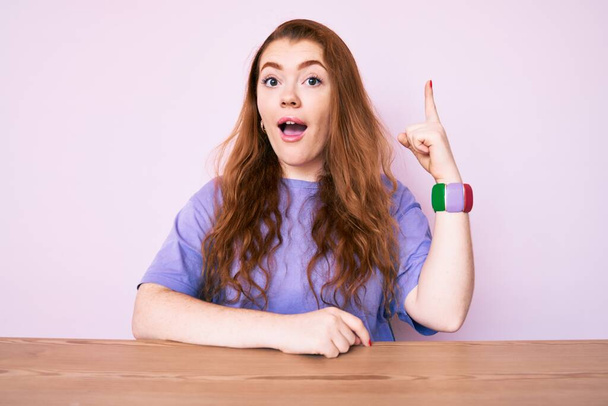 Young redhead woman wearing casual clothes sitting on the table smiling with an idea or question pointing finger up with happy face, number one  - Photo, image