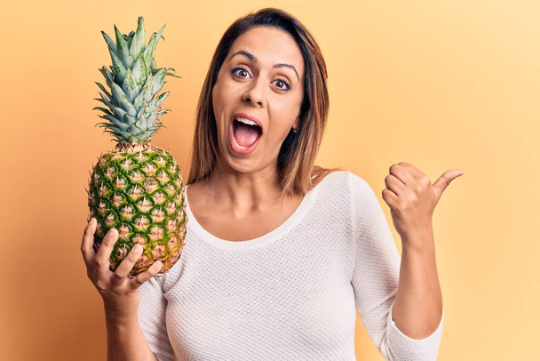 Nuori kaunis nainen, jolla ananas osoittaa peukalolla sivulle hymyillen onnellisena avoimella suulla  - Valokuva, kuva