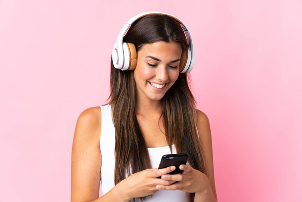 Νεαρή Βραζιλιάνα κοπέλα απομονωμένη σε ροζ φόντο ακούγοντας μουσική και κοιτώντας στο κινητό - Φωτογραφία, εικόνα