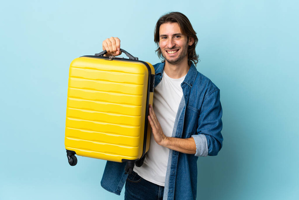 Mladý pohledný muž izolovaný na modrém pozadí v dovolené s cestovní kufr - Fotografie, Obrázek