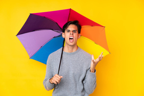 Homme tenant un parapluie sur fond jaune isolé malheureux et frustré par quelque chose - Photo, image