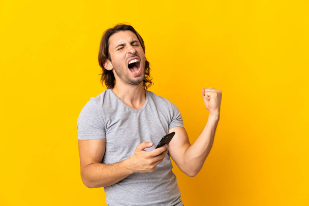 Junger gutaussehender Mann isoliert auf gelbem Hintergrund mit Handy in Siegesposition - Foto, Bild