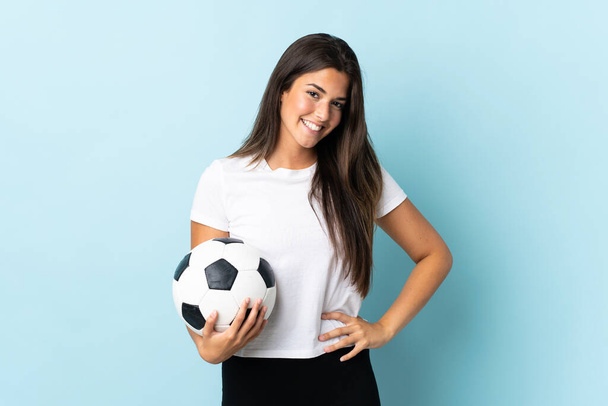 Junge Fußballerin brasilianisches Mädchen isoliert auf blauem Hintergrund posiert mit Armen an der Hüfte und lächelt - Foto, Bild