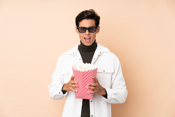 Człowiek na odosobnionym tle zaskoczony okularami 3D i trzymając duże wiadro popcornów - Zdjęcie, obraz