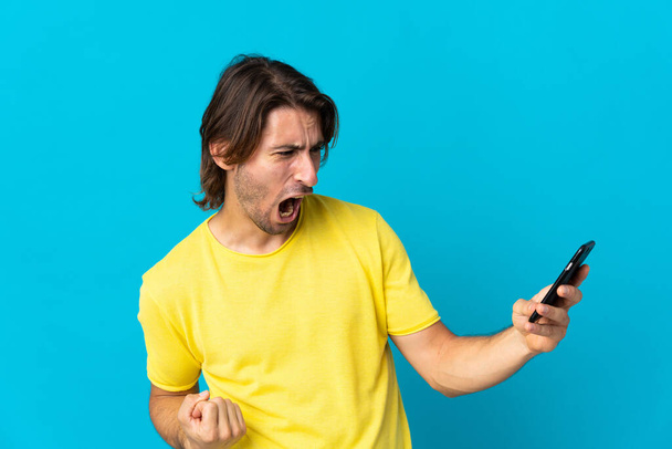 Mladý pohledný muž izolovaný na modrém pozadí pomocí mobilního telefonu a dělá vítězný gesto - Fotografie, Obrázek