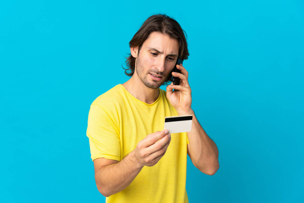 Jeune bel homme isolé sur fond bleu acheter avec le mobile avec une carte de crédit - Photo, image