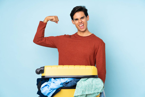 Мужчина с чемоданом, полным одежды на изолированном синем фоне делает сильный жест - Фото, изображение