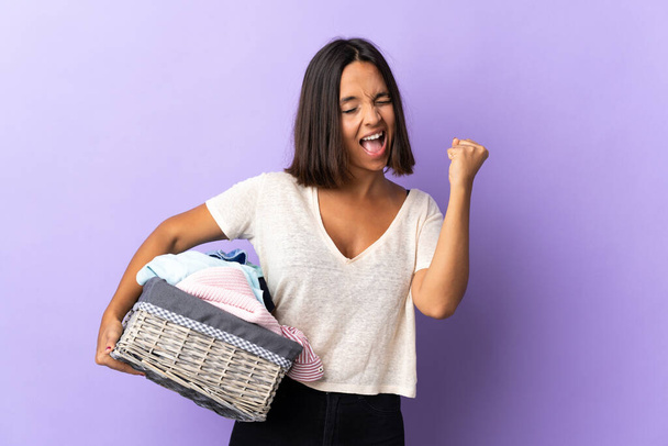 Young latin woman holding a clothes basket isolated on purple background celebrating a victory - Φωτογραφία, εικόνα