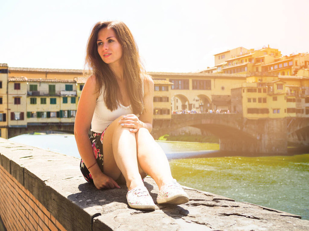 Une belle jeune femme en jupe est assise sur le remblai de Florence sur fond de célèbre pont - Photo, image
