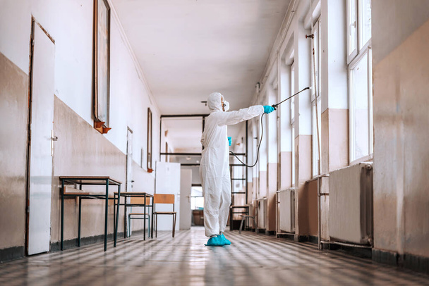 Worker in sterile white uniform, with mask and glasses holding sprayer with disinfectant and spraying around hallway in school. Prevention of spreading corona virus. - Photo, Image