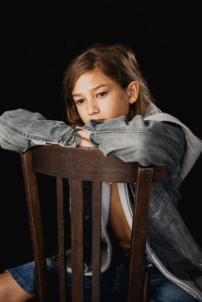 Eleven years old boy with hooded sweater and a jean jacket sitting on a wood chair against a black background - Foto, Imagen