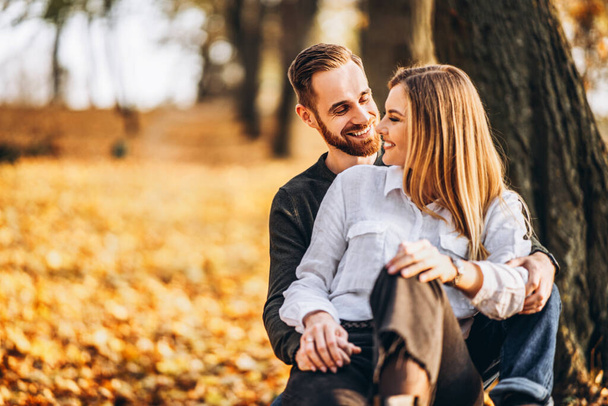 Een jong liefdevol stel zittend op een houten bank in het bos. man en vrouw knuffelen en glimlachen op de achtergrond van de herfst bomen. - Foto, afbeelding
