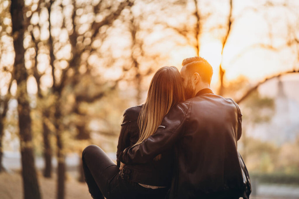 Ein romantisches Paar, das auf einer Bank sitzt, sich umarmt und den Sonnenuntergang im Park beobachtet. Liebesgeschichte. Rückseite. - Foto, Bild