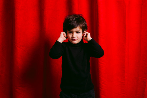 Τρία χρόνια αγόρι, φορώντας μαύρα ρούχα, καλύπτοντας τα αυτιά του, πάνω σε μια κόκκινη κουρτίνα - Φωτογραφία, εικόνα