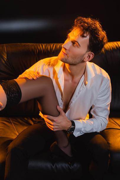 страстный мужчина в рубашке сидит на диване рядом с женщиной в чулке на черном  - Фото, изображение