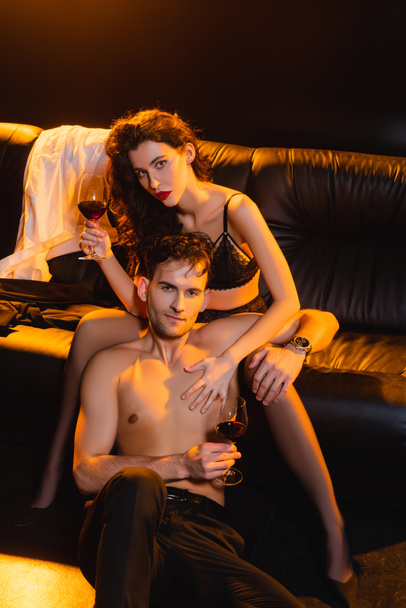 muscular man and seductive woman holding glasses of red wine while looking at camera on black  - 写真・画像