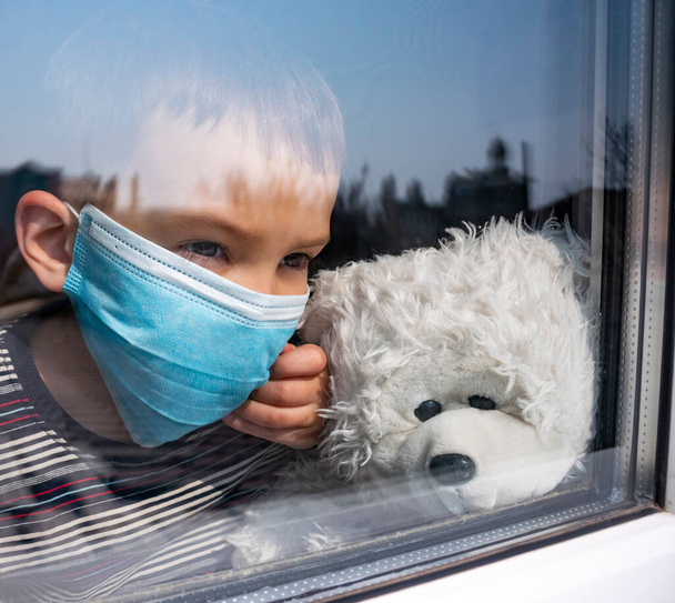 Маленький хлопчик з плюшевим ведмедем носить захисну медичну маску, яка дивиться крізь вікно. Карантин коронавірусу під час пандемії COVID-19. Залишайтеся вдома
. - Фото, зображення