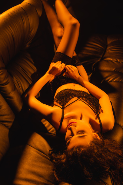 young woman with closed eyes in lace underwear lying on black sofa - Foto, afbeelding