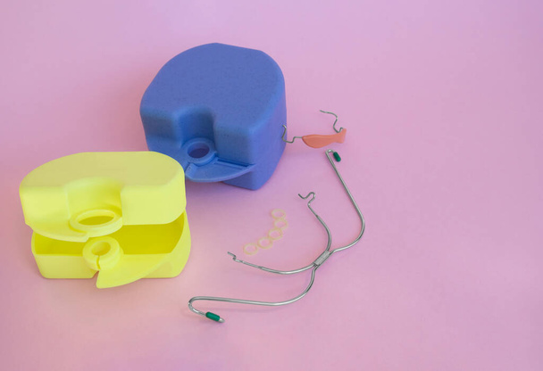 歯列矯正装置:取り外し可能な金属製のブレースと上下の歯のためのゴム製の肉色のプラスチックキャップ、歯科用ゴムバンド、ピンクの背景に黄色と青のボックス。 - 写真・画像