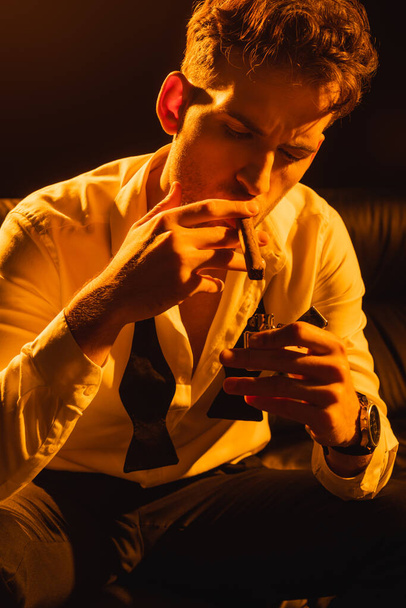 man in suit holding lighter near cigar while sitting on sofa on black  - Foto, Bild