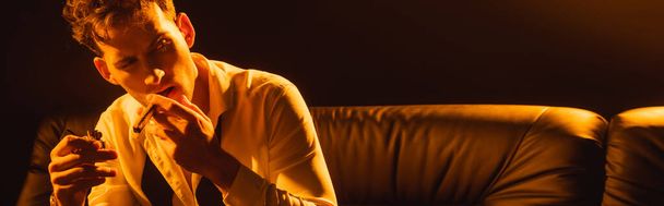 cultivo panorámico del hombre en traje sosteniendo más ligero, fumar cigarro, sentado en el sofá y mirando hacia otro lado en negro  - Foto, Imagen