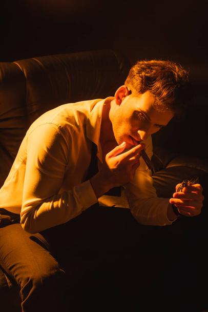 man in suit holding lighter, smoking cigar and sitting on sofa isolated on black  - Φωτογραφία, εικόνα