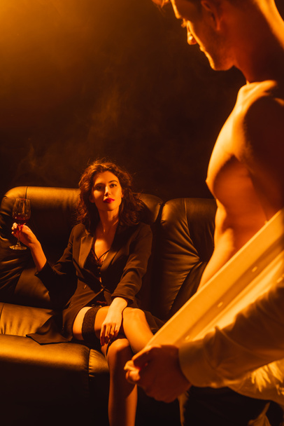 selectieve focus van vrouw met glas rode wijn zittend op de bank en kijkend naar gespierde man op zwart met rook - Foto, afbeelding