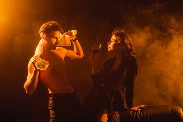 mulher sedutora em blazer segurando copo de vinho tinto enquanto olha para o homem muscular em preto com fumaça - Foto, Imagem