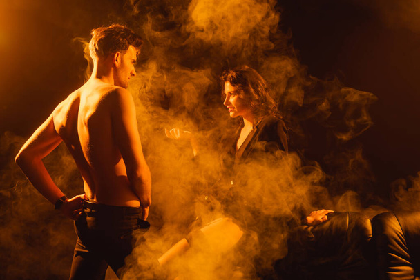 curly woman in jacket looking at shirtless man standing with hand on hip on black with smoke  - Zdjęcie, obraz