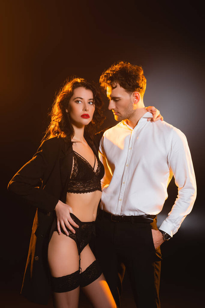 man in suit standing with hand in pocket and looking at seductive woman in underwear and jacket on black  - Foto, imagen
