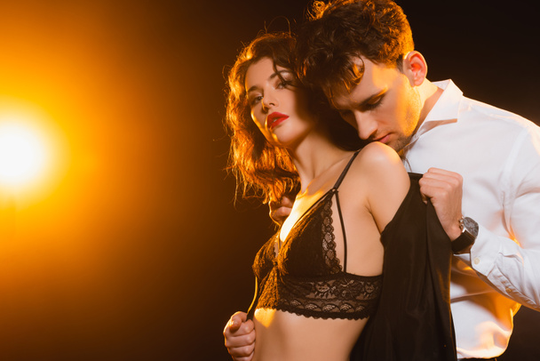 passionate man in suit taking off jacket on sexy woman in bra on black with back light  - Photo, Image
