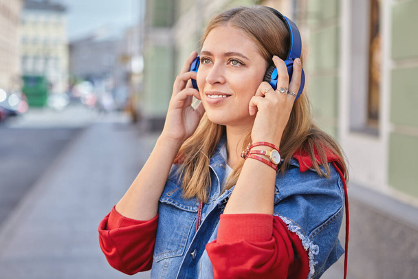 Young attractive caucasian woman with blond hair and fashionable cloth about 25 years old is standing outdoor and listening to music with help of earphones. - Valokuva, kuva