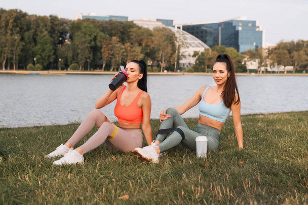 As mulheres jovens relaxam depois de um treino de fitness ao ar livre. Uma menina está cansada e bebe água
 - Foto, Imagem