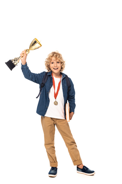 colegial rubia con medalla de oro sosteniendo trofeo y libro aislado en blanco  - Foto, Imagen