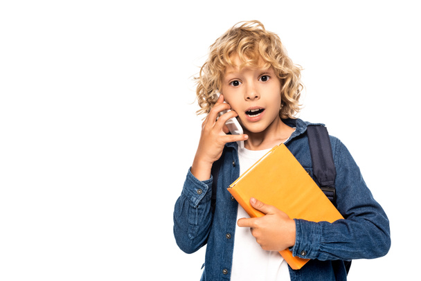 surprised blonde schoolboy holding book and talking on smartphone isolated on white  - Photo, image