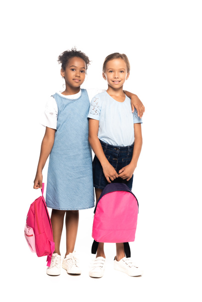 multicultural schoolgirls holding backpacks while hugging isolated on white  - Fotó, kép