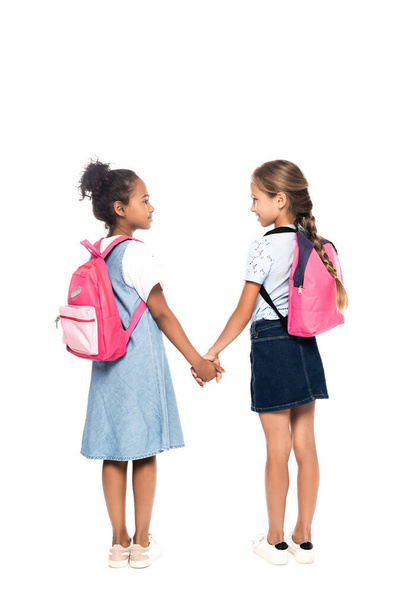 multicultural schoolkids with backpacks holding hands and looking at each other isolated on white  - Foto, imagen