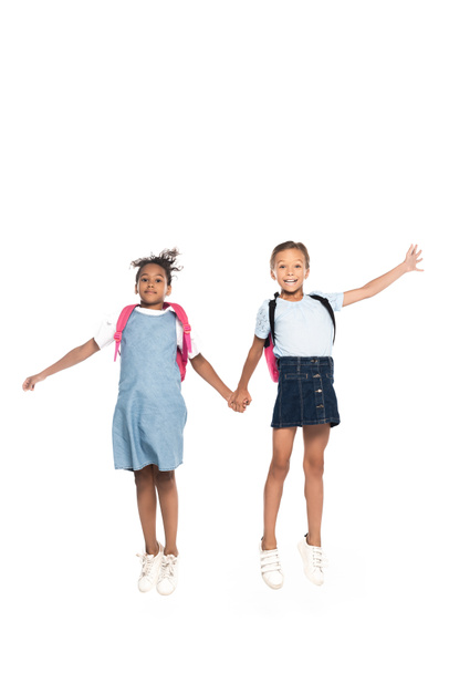multicultural schoolgirls with backpacks holding hands and jumping isolated on white  - Foto, imagen