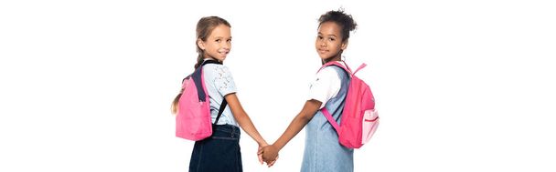 горизонтальное изображение одноклассников с рюкзаками, держащимися за руки и смотрящими на камеру, изолированную на белом  - Фото, изображение