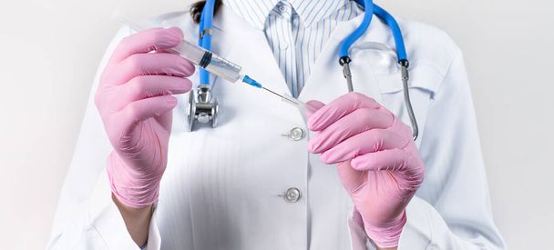 Gewassen zicht op jonge vrouwelijke arts in een witte jas en roze steriele handschoenen houden de spuit vast. Focus op de spuit. Inentingsconcept. - Foto, afbeelding